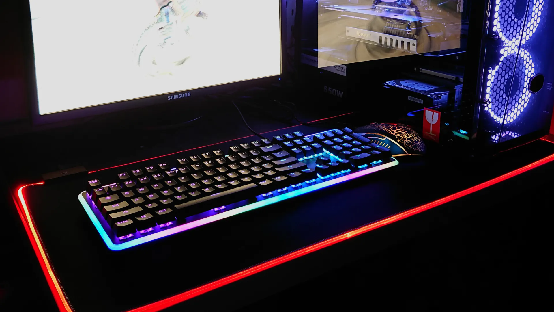 An LED illuminated gaming keyboard stands on a gaming table next to a gaming PC with LED components.
