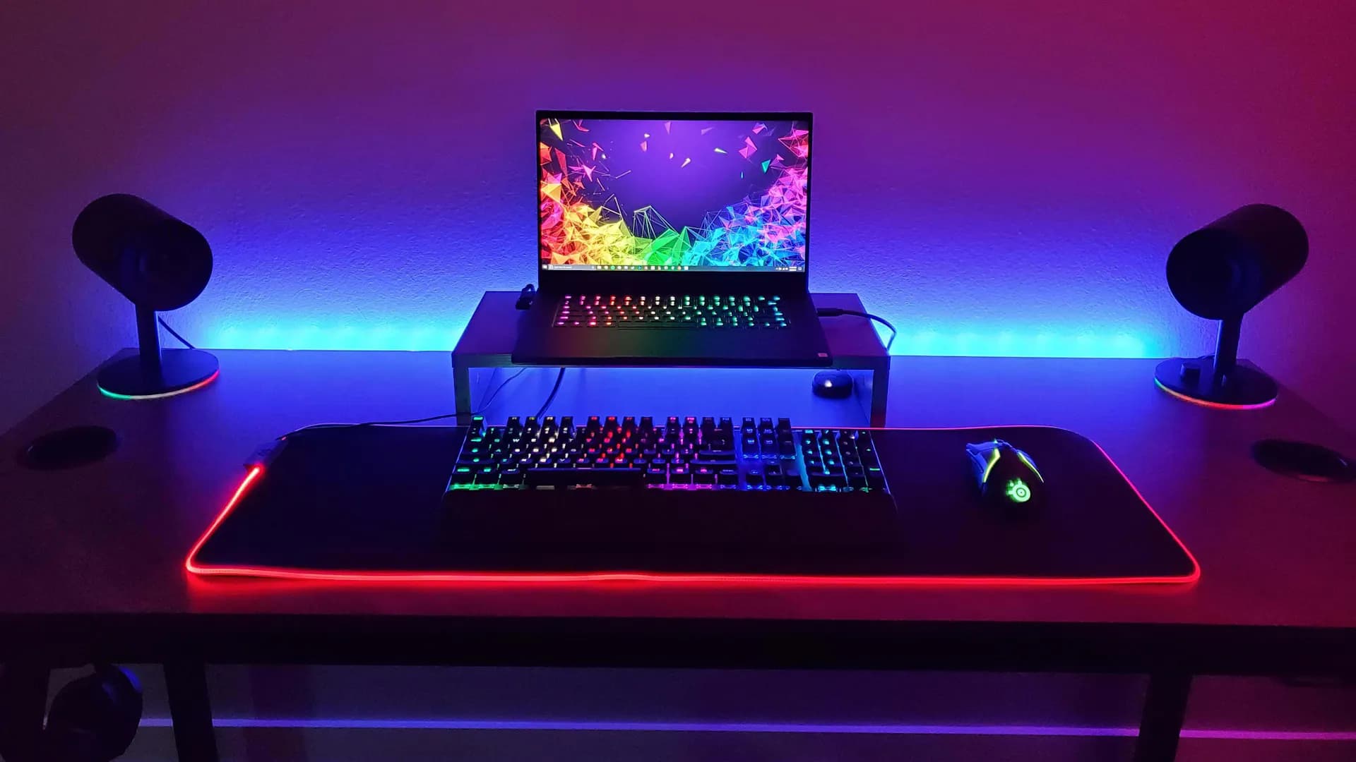 On a table without decoration but with many colorful gaming LED elements, a laptop stands frontally somewhat elevated behind a gaming keyboard.